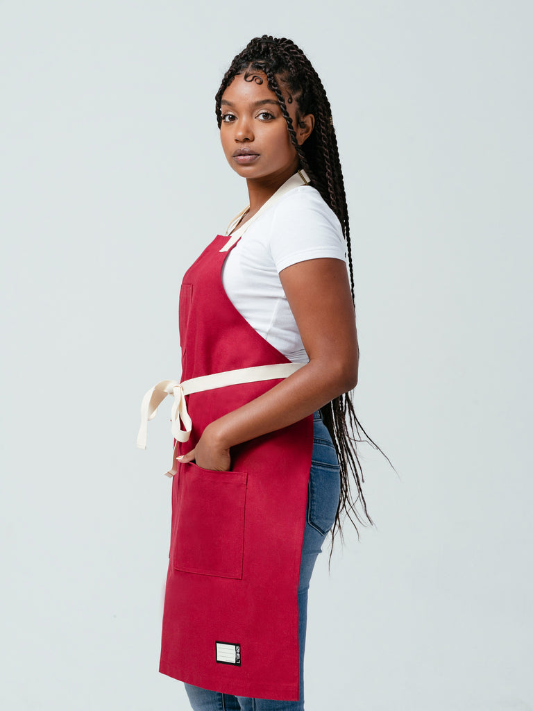 Woman looking at camera with hands in her pockets modeling Helt's Burgundy Red Bib Apron.