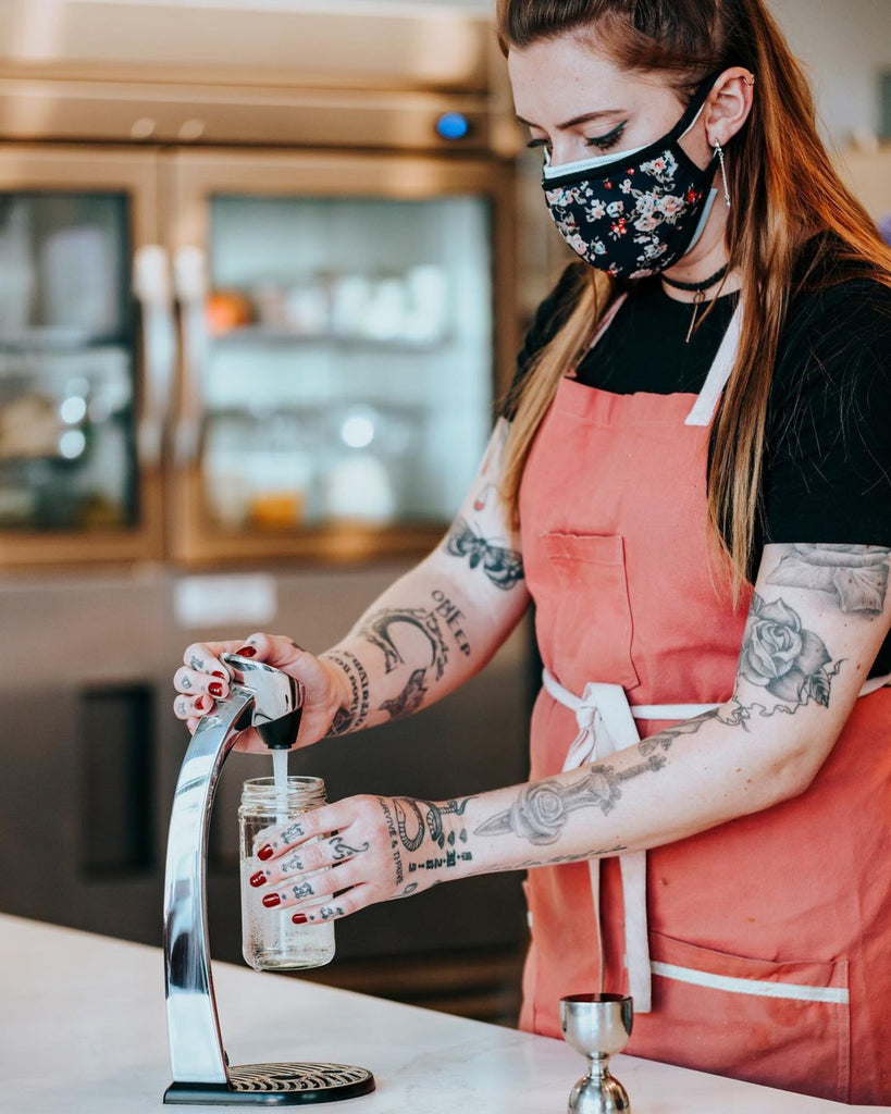 Tattooed and masked female bartender making a drink and wearing bib apron.