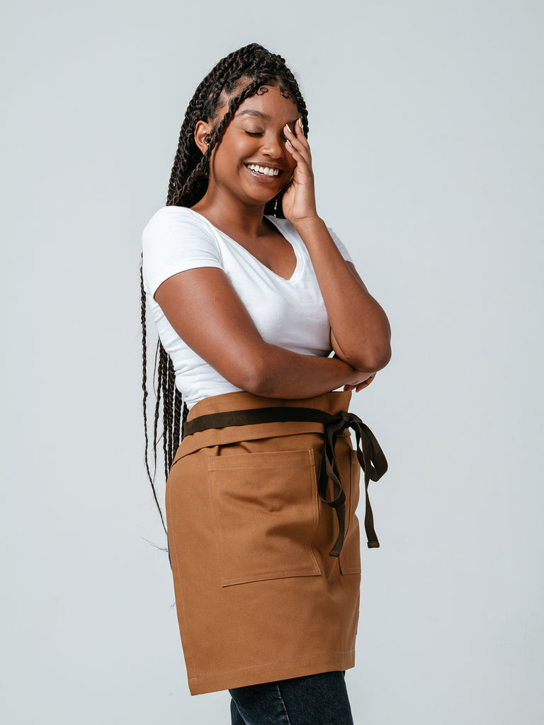 Woman with hand on her face, smiling, and modeling Helt Studio's Ranch Tan DWR Bistro Apron.
