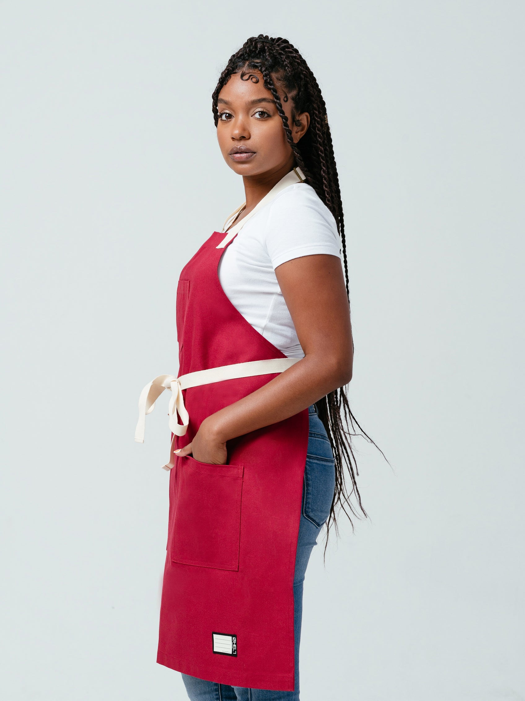 Woman looking at camera with hands in her pockets modeling Helt's Burgundy Red Bib Apron.