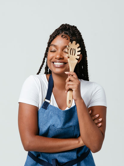 Woman holding wooden pasta fork over eye, laughing, modeling Helt's Ocean Breeze Denim Apron.
