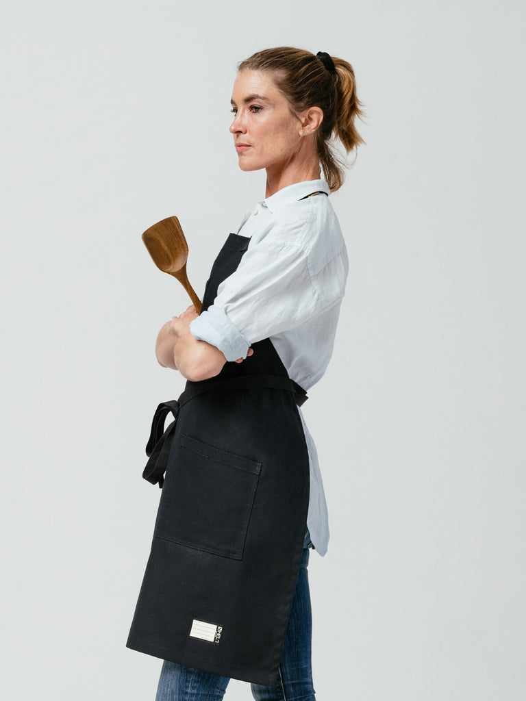 Side-view of woman holding wooden spatula modeling Helt's Raven Canvas Apron.