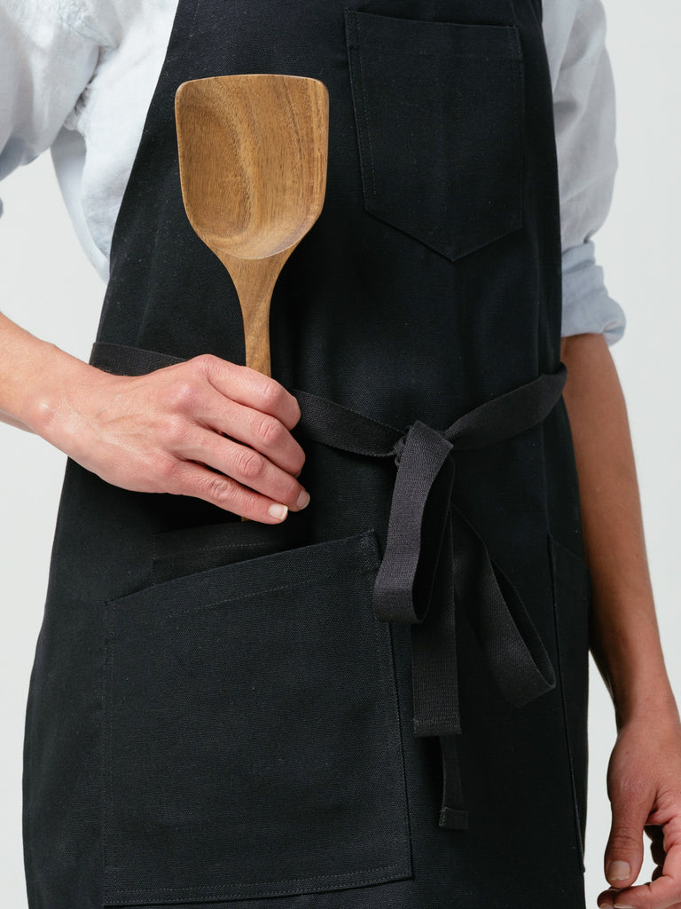 Close up of woman placing wooden spatula in pocket of Helt Studio's Raven Canvas Apron.