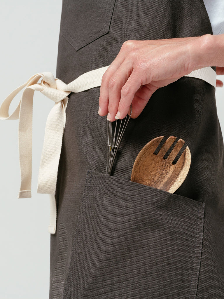 Close up of woman placing utensils in pocket of Helt Studio's Hardwood Briquette Bib Apron.