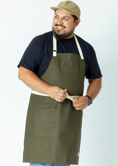 A person wearing an olive green gardening apron with pockets, standing against a white background.