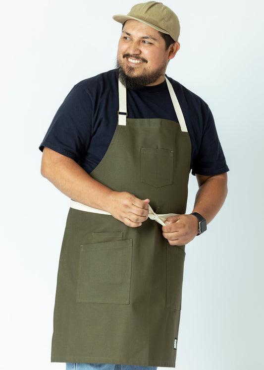 A person wearing an olive green gardening apron with pockets, standing against a white background.